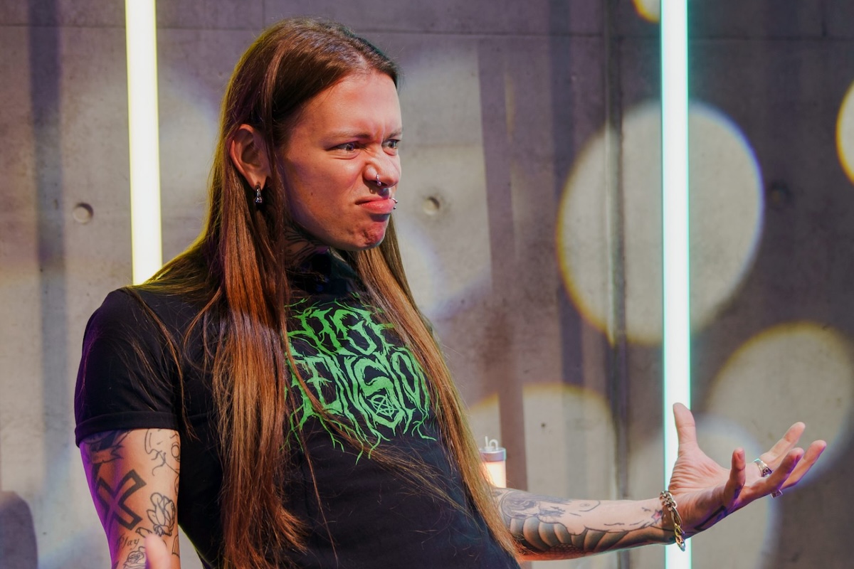 Australian comedian Nat's What I Reckon making an animated face on stage, wearing a green graphic metal band t-shirt with long hair and tattoos.